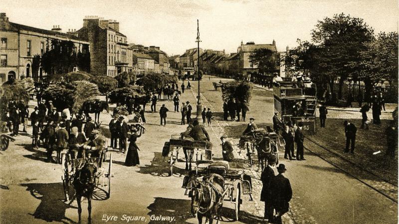 The Hay Market in Eyre Square, Galway in the early years of the 1900s. Note the tram in the middle of the road, the railings around the park and the barefooted children in the centre of the photograph.