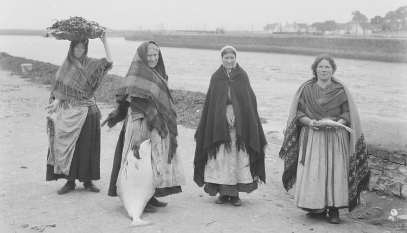 Women from the Claddagh (in background) wearing their traditional shawls at Fishmarket (now Spanish Parade) in Galway in 1905. Seaweed (being carried by the woman on left) is stacked along the quay wall, while the woman on second left is holding an impressive large flatfish of some sort.