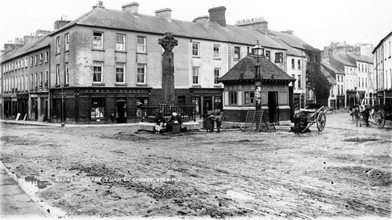 Market Square in Tuam in the late 19th Century. The photo was possibly taken in the 1890s. Three ladies in shawls typical of the time are seated at the base of the High Cross of Tuam while three children play beside them. Behind, a barefoot boy stands on the footpath outside James J Begley's general store. To the right of that is the drapery of M Nolan. The 12th Century High Cross of Tuam was moved from this location to St Mary's Cathedral in 1992.
